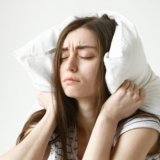 pretty-student-woman-in-striped-t-shirt-trying-to-have-some-sleep-sitting-in-bedroom-with-pillow-on-her-head-closing-eyes