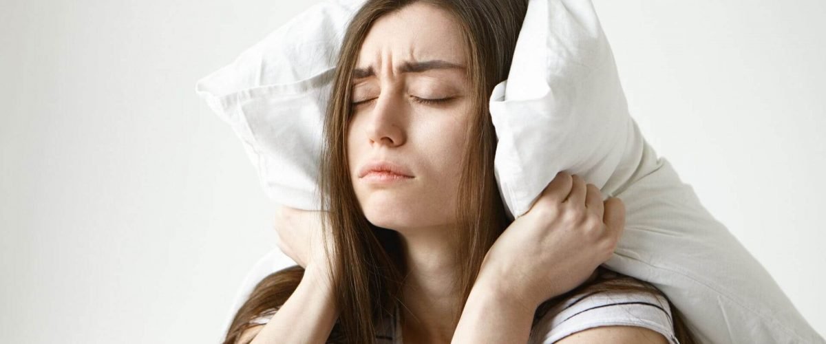 pretty-student-woman-in-striped-t-shirt-trying-to-have-some-sleep-sitting-in-bedroom-with-pillow-on-her-head-closing-eyes